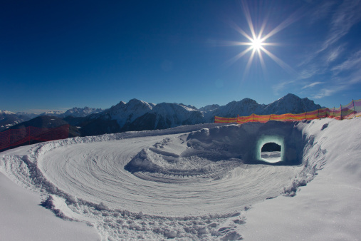Winter panorama showing a ski slope going through the tunnel made out of snow with mountains in background and sun rays.