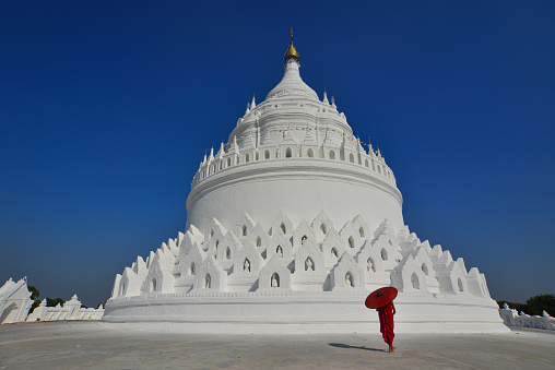 A Burmese Buddhist novice monk walking around the white stupa in sunny day.