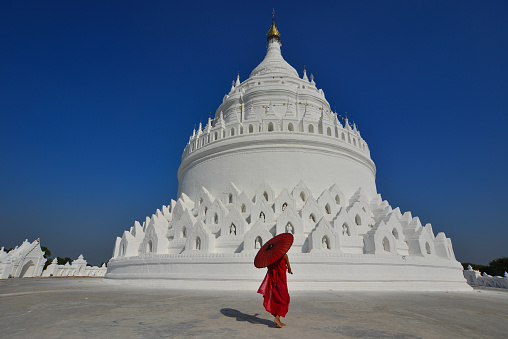A Burmese Buddhist novice monk walking around the white stupa in sunny day.