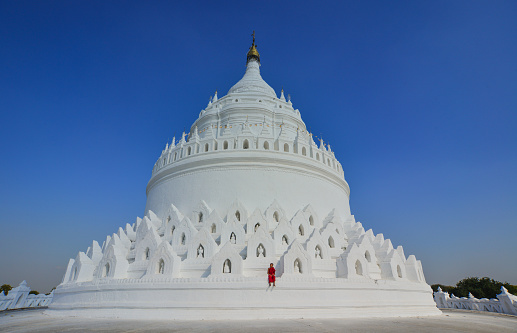 A Buddhist novice monk with a red umbrella walking around the white stupa in sunny day.