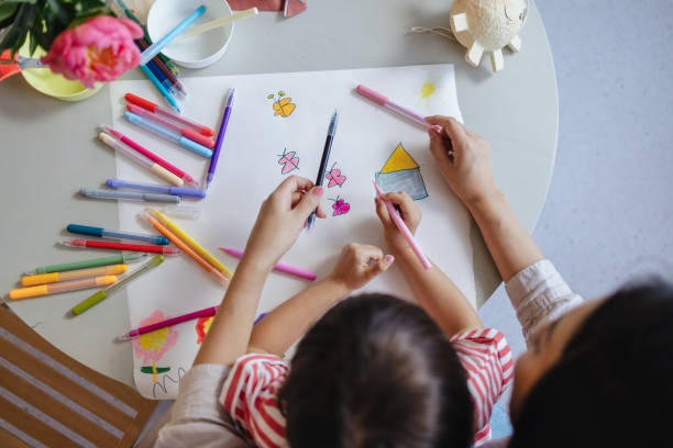 A From Above View Of An Unrecognizable Cute Asian Girl And Her Unrecognizable Nanny Drawing Using Markers A close up view of an unrecognizable preschool Japanese child developing her creativity while sitting at the table, in her mother's lap. asian nanny stock pictures, royalty-free photos & images