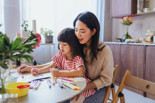 A Happy Cute Asian Girl Drawing With Her Nanny Using Markers An adorable preschool Japanese child developing her creativity while sitting at the kitchen table with her smiling mother. parent and kid stock pictures, royalty-free photos & images
