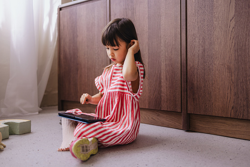 A wide angle view of a serious adorable playful Japanese child using tablet while sitting on the floor at home.