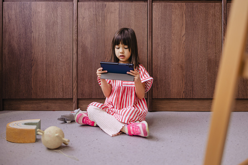 A serious adorable playful Japanese child using tablet while sitting on the floor at home.