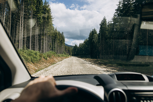 From the vantage point of the driver's seat, a car navigates a winding dirt road that meanders through a picturesque forest nestled within the embrace of majestic mountains. The steering wheel rests comfortably in the driver's hands as they skillfully guide the vehicle along the rugged path.