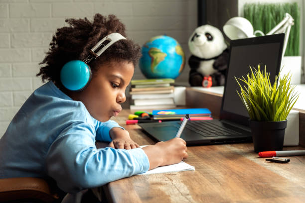 African-American girl doing homework,making notes,using headphones and laptop at home.Back to school concept.School distance education at home,home schooling,e-learning,diverse people