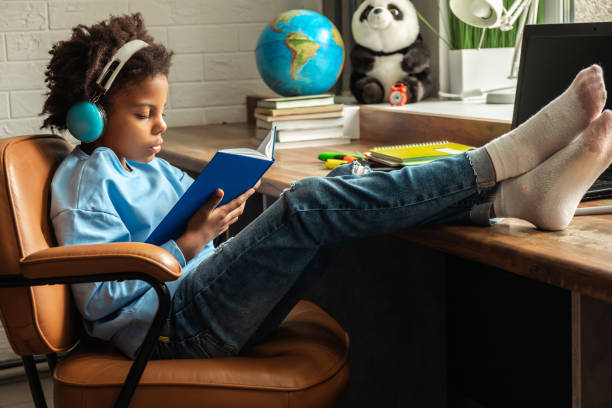 African-American girl doing homework,reading a book,using headphones and laptop at home.Back to school concept.School distance education at home,home schooling,e-learning,diverse people