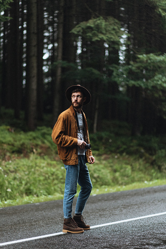 Amidst the striking mountainous terrain, a fashionable and attractive male photographer, sporting a hat and carrying his camera, stands amidst a backdrop of captivating forest scenery.