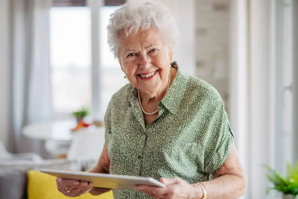 Portrait of beautiful senior lady with tablet in hands at home. Happy elderly woman uses tablet for shopping, making video calls, reading newspaper or playing card games.