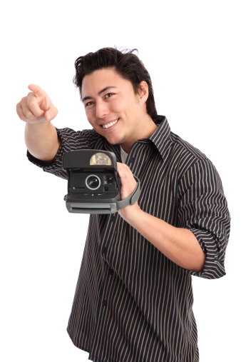 Young man pointing, holding a polaroid camera. Wearing a striped shirt. White background.