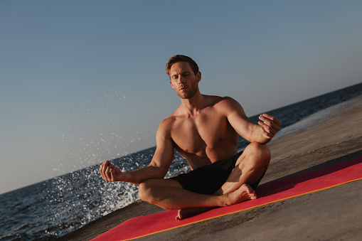Confident young shirtless man sitting on exercise mat while meditating seaside