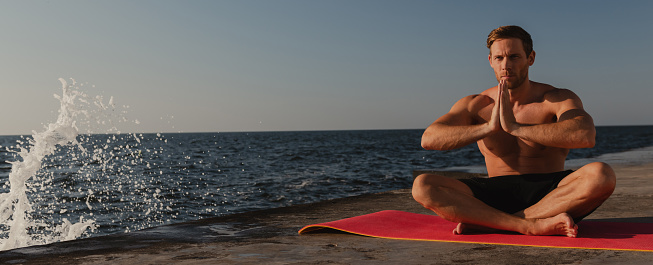 Confident young shirtless man practicing yoga while meditating near the sea at the morning