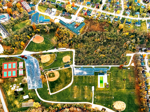 A drone view of a neighborhood sporting events venue between Dayton and Cincinnati Ohio. The complex features practice fields, soccer, baseball, and softball fields. There are basketball, pickle-ball and tennis courts.  There is a lap pool. There is a walking trail. There are parking lots. You can seen the surrounding housing and business developments.