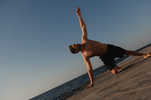 Handsome young shirtless man practicing yoga outdoors with the sea on background