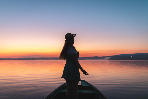 Silhouette of a happy young woman standing on a boat in the middle of a lake.