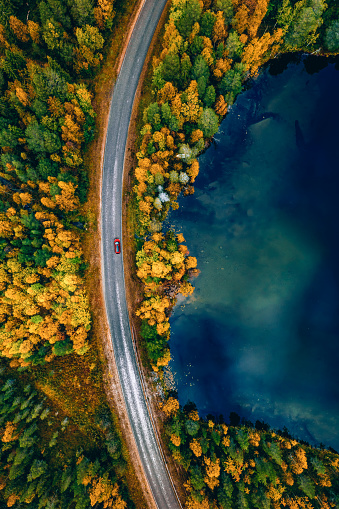 Aerial view of road with red car by blue lake and fall forest with autumn colorful trees. Beautiful nature landscape in Finland