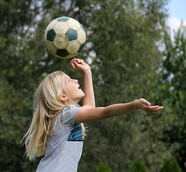 This is a photo of a teenage female athlete holding a ball on a beach volleyball court in a park setting. This photo is taken from a low angle capturing a great cloudscape behind her. She is looking tough and competitive out into the horizon.