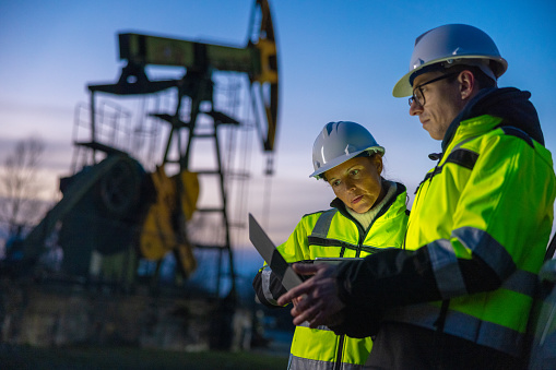 Male and female professionals in neon vests and hardhat planning over laptop while standing against oil pump on field during sunset