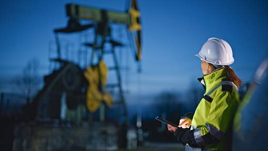 Female engineer in protective workwear looking at machinery while using tablet PC at oil field against blue sky during dusk