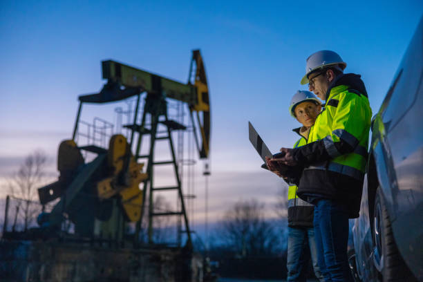 Partners planning over laptop while standing against equipment by car at field Low angle view of male and female partners in protective clothing planning over laptop while standing against equipment by car at field against sky during sunset drill team stock pictures, royalty-free photos & images