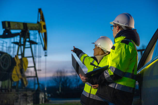Engineers looking at machinery while planning over laptop by car at oil field stock photo