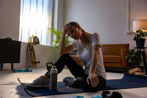 Tired female athlete sitting with head in hand on exercise mat after workout at home