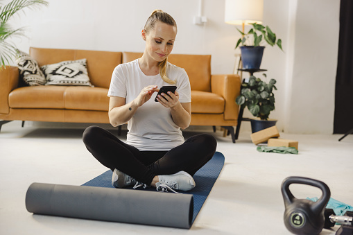 Smiling young sportswoman using smart phone after workout while sitting on exercise mat at home