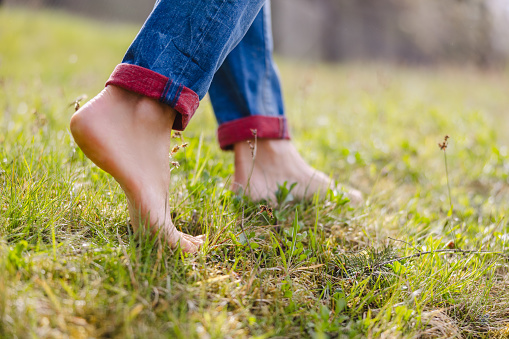Close-up low section of woman in jeans walking with barefoot on grassy field at park during sunny day