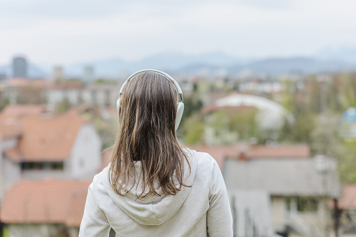 Rear view of woman with brown hair listening music through headphones while looking at houses in town against sky