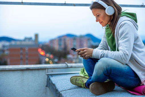 Female wearing sweater using smart phone and enjoying music through headphones while sitting cross-legged at terrace during sunset