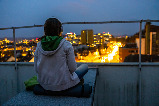 Rear view of woman listening music through headphones while sitting on terrace in illuminated city against sky at dusk