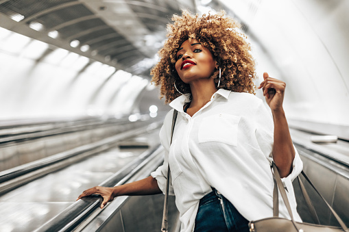 Beautiful black woman standing on escalator on her way to modern brightly lit subway station. Public transportation and urban life concept. Low angle shot.