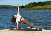 Woman practicing yoga doing Utthita Parshvakonasana exercise, extended side angle pose, exercising in sportswear on the lakeside on a warm sunny morning