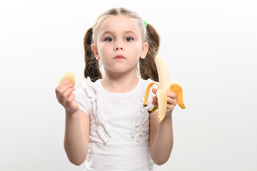 Portrait of a girl in the studio on a white background, eating a banana and holding chips in her hand, the concept of healthy and unhealthy food.