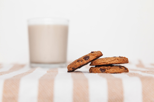 Close-up of stacked cookies with chocolate drops and a glass of milk over a striped cloth mat in a table. Selective focus on pile of cookies, white background.