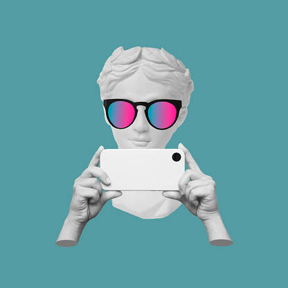 Antique statue's head in sunglasses holding mobile phone with photo camera in hands taking picture on color background. 3d trendy creative collage in magazine style. Contemporary art. Modern design