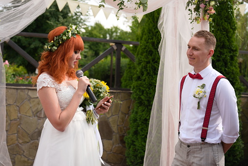red-haired bride with flowers reads from the phone into the microphone to the groom in a white shirt and red suspenders. Ukrainian wedding on the street in boho style.