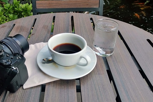 A veiw of a black coffee cup on wooden table with camera and glass of water with a garden background.
