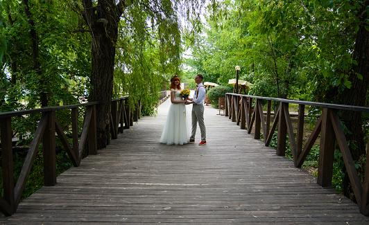 Young happy bride and groom hold hands and look at each other on the bridge under green foliage. Happy boyfriend and girlfriend, young family, newlyweds.