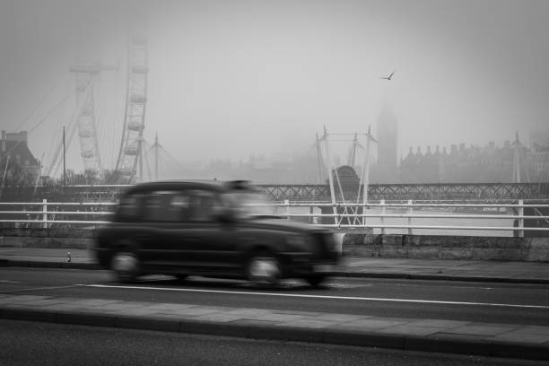 A black and white photograph of a traditional London taxi cab that is driving across a bridge with the iconic London eye wheel in the background.