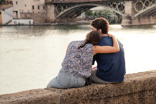 Seville, Andalusia, Spain; June 4th 2023: Lovers sitting by the Guadalquivir River in Seville. A romantic scene where a girl embraces her boyfriend. A display of affection in a couple.