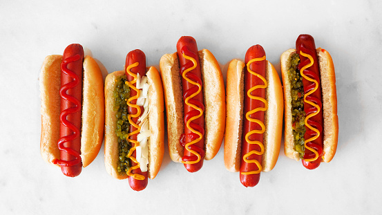 Variety of unique hot dogs with an assortment of toppings. Overhead view on a white marble background.