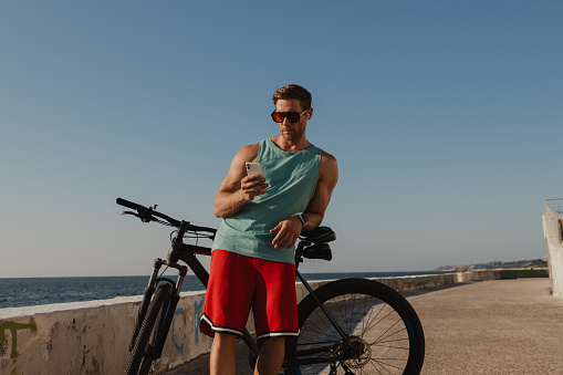 Handsome young man in headphones using smart phone while leaning on his bicycle seaside