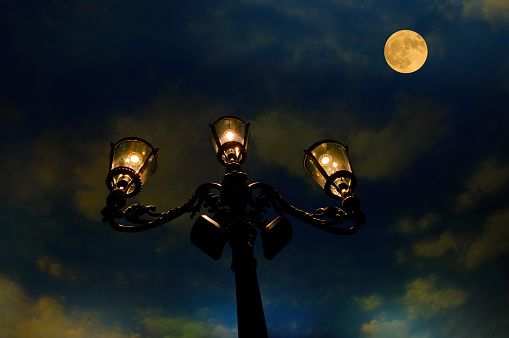 Vintage, ornate wrought iron street lamps at dusk against a bright, colorful, yellow full moon.
