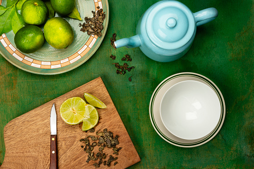 High angle vie of table with healthy ingredients to drink a fresh healthy green tea at home.Empty bowl, cutting board with slices of lemon and dry green tea leaves and a blue pot with hot water.