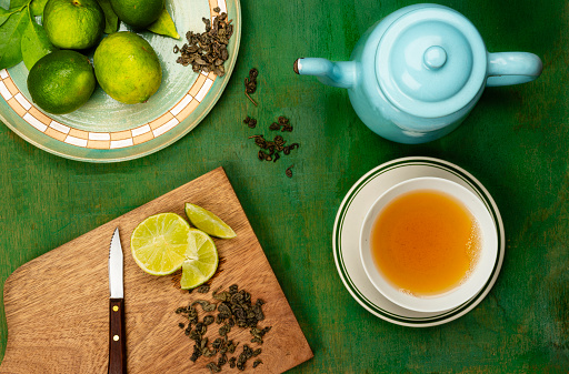 Tea time with healthy green tea.\nHigh angle view of bowl with green tea. In the image you can see a cutting board with slice of lemon, dry green tea leaves, and a blue pot with hot water