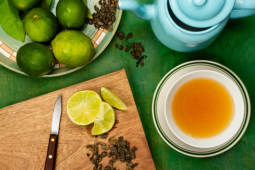 Tea time with healthy green tea.
High angle view of bowl with green tea. In the image you can see a cutting board with slice of lemon, dry green tea leaves, and a blue pot with hot water