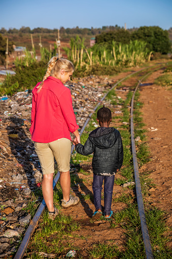 African little girl walking with young caucasian woman on railroad tracks, Kibera slum on the background, Kenya, East Africa. Kibera is the largest slum in Nairobi, the largest urban slum in Africa, and the third largest in the world