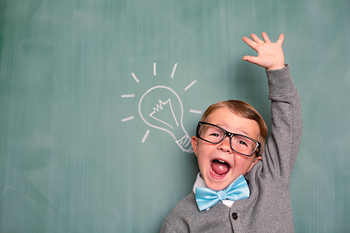 A young boy dressed in retro attire is full of new and inventive ideas and is standing in front of a light bulb drawn on a chalkboard.