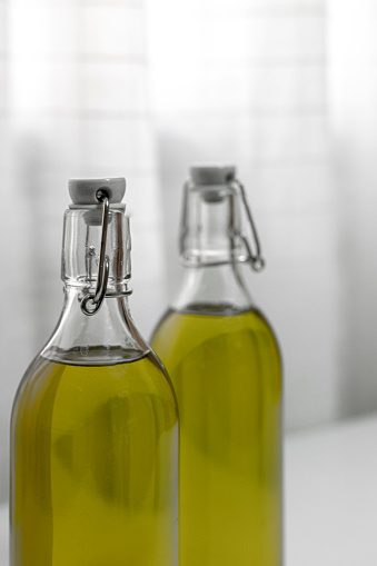 Extra virgin olive oil is bottled in glass with hermetic cap, with an olive green color, on the countertop of a kitchen table, there are fabric curtains behind it, with a white background.